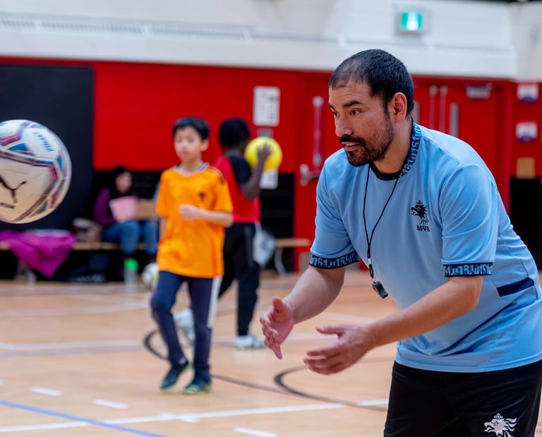 A youth soccer coach in a blue jersey training children during an indoor soccer sports class at MVB