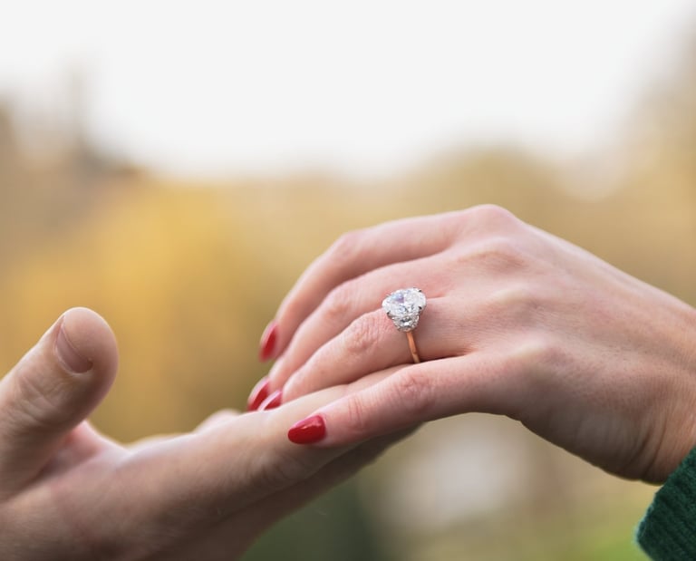 gros plan sur un main d'homme soulevant la main d'une femme portant une bague de fiançailles, le fond est lumineux et flou.