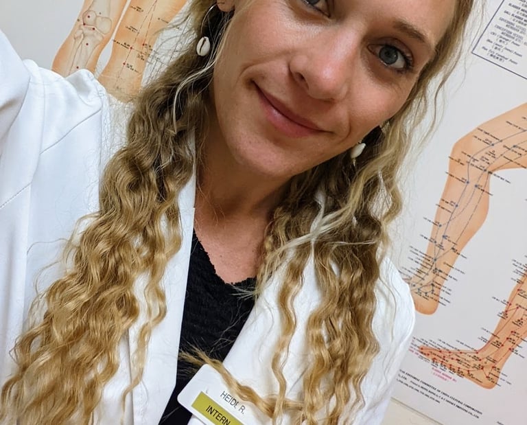 blonde acupuncture intern in a white lab coat posing in front of anatomical meridian charts.