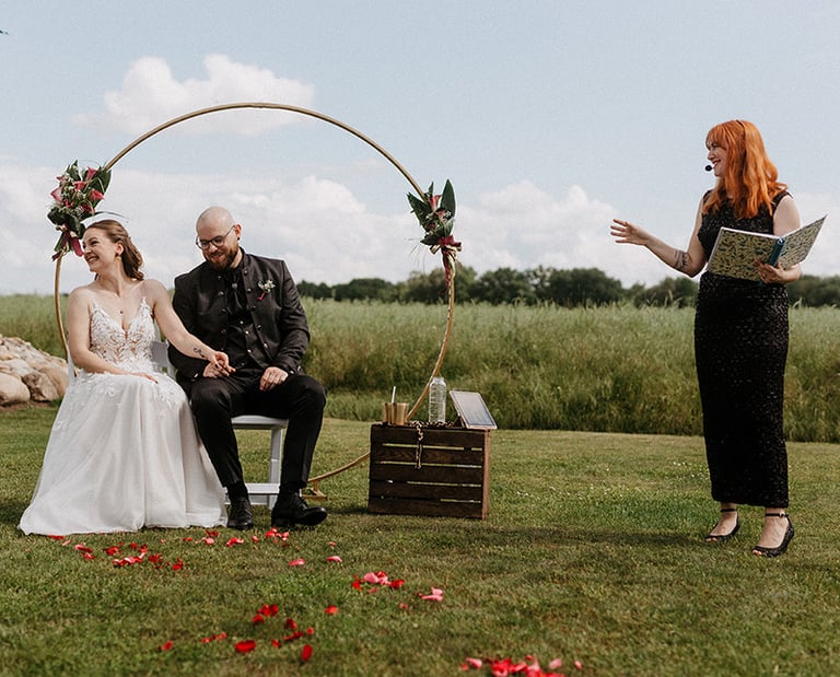grinsend sitzen Braut und Bräutigam nebeneinander während die Traurednerin im schwarzen Glitzerkleid eine Anekdote erzählt