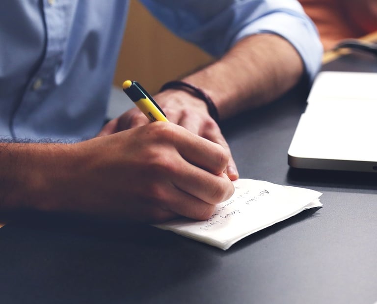 A person writing notes with a yellow pen on paper next to a laptop on a desk.