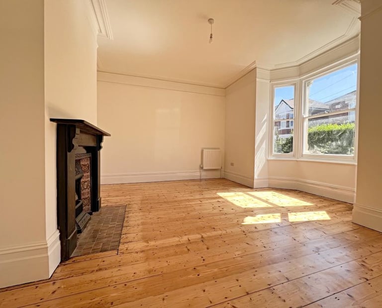 Spacious empty living room featuring original pine wood flooring, a vintage fireplace, and a bright bay window.