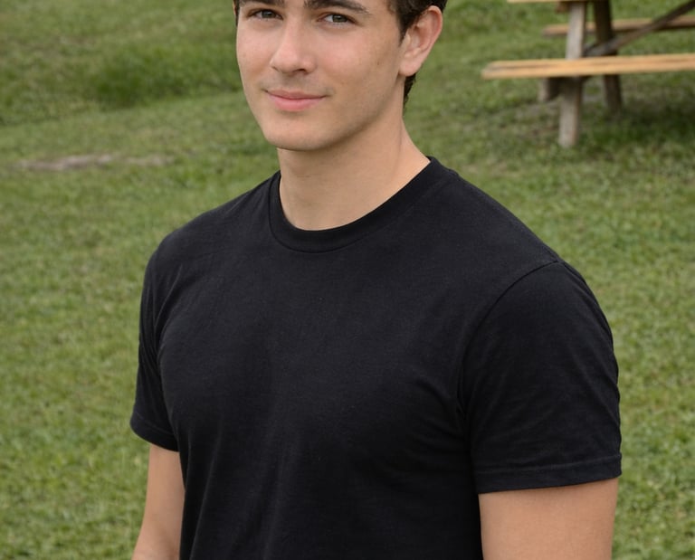Young man with brown curly hair wearing a black t-shirt outdoors at a park with picnic tables.