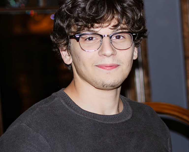 Young man with curly brown hair and glasses wearing a dark sweater in a portrait.