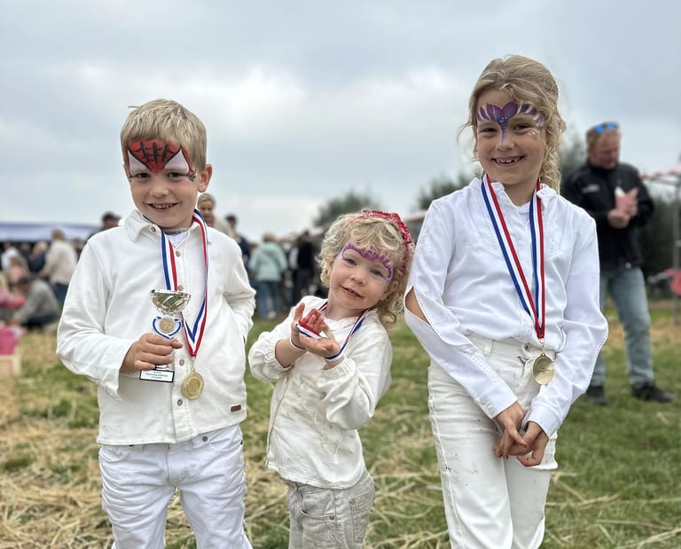 Drie trotse kinderen met medailles en schminkcreaties op een evenementenveld.