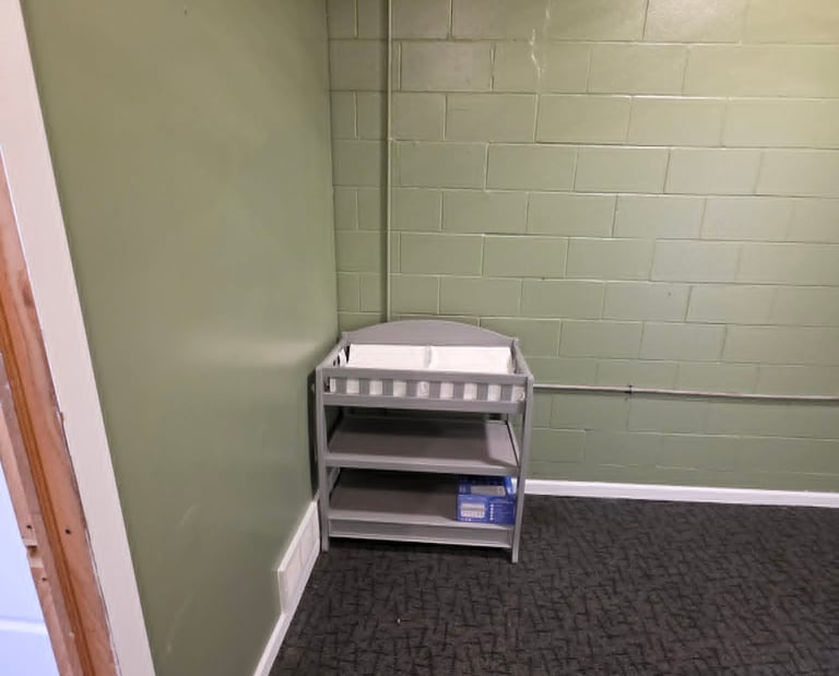 A gray wooden baby changing table with shelves stands against a green cinder block wall in a nursery room.