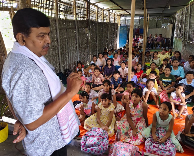 An educator teaching a large group of attentive children sitting on the floor in a bamboo-walled classroom.
