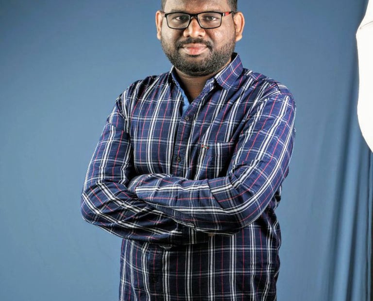 Professional portrait of a man in a plaid shirt with arms crossed against a blue backdrop.