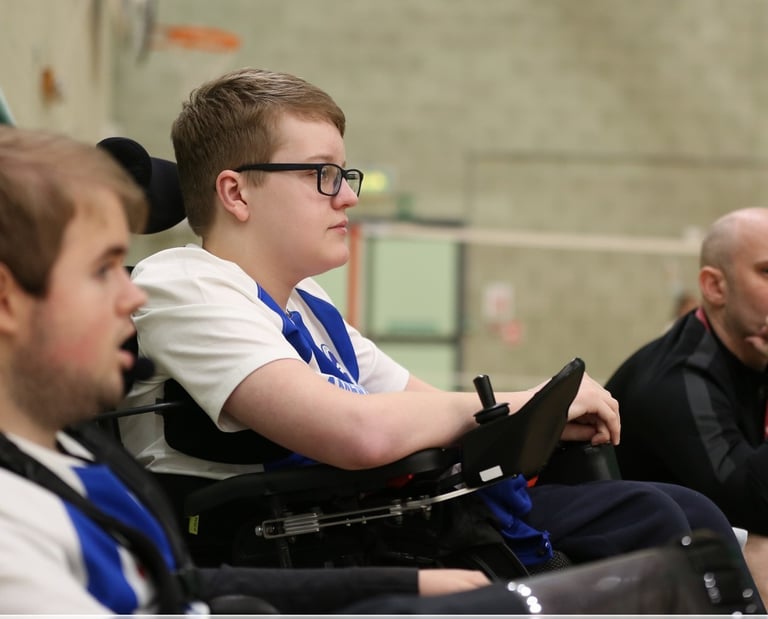 Indoor sports setting with young powerchair football players sitting side-by-side and watching