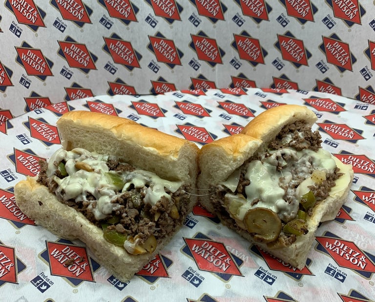 A sliced Philly cheesesteak sandwich with melted white cheese and peppers on Dietz & Watson paper.