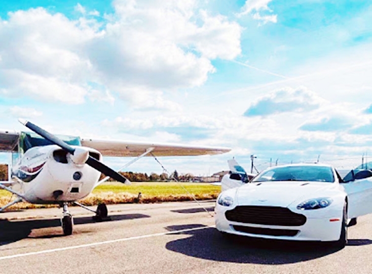 A white Aston Martin luxury sports car parked next to a small private propeller plane on an airfield runway.