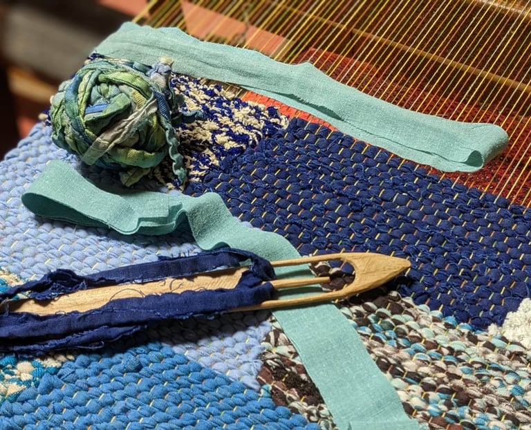 a weaving loom with a wooden shuttle, a ball of rag threads and a rag rug in shades of blue in progr