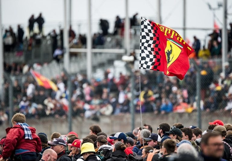 Ferrari flag waving over a crowded grandstand during a Formula 1 racing event at a motorsports track.