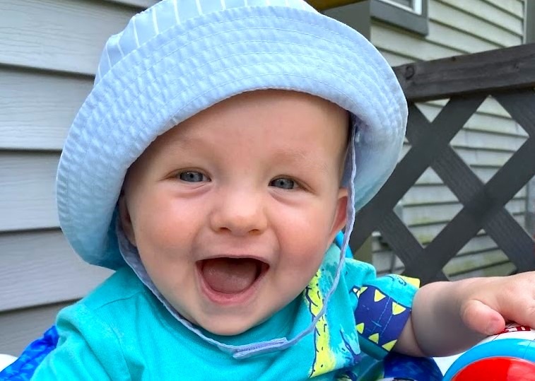 A happy smiling baby boy wearing a light blue sun hat and a dinosaur t-shirt outdoors.