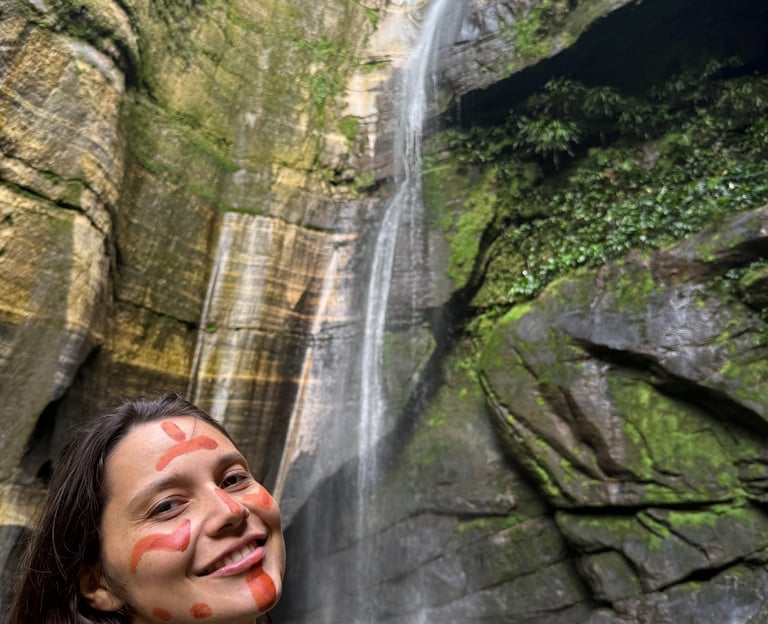 Une femme souriante, le visage peint selon la tradition indigène, se tient devant une haute cascade