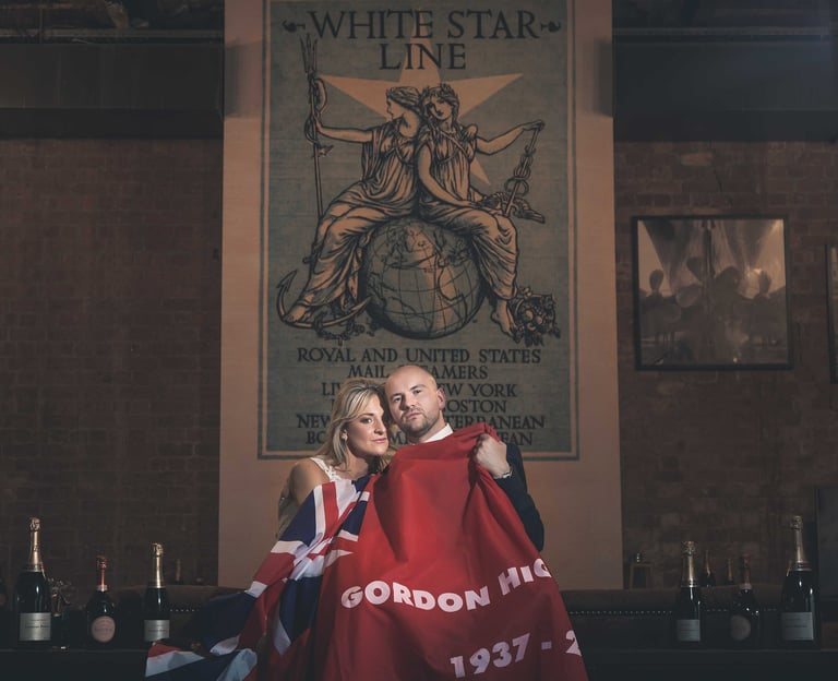 Couple holding a Union Jack flag under a vintage White Star Line poster in a brick room.