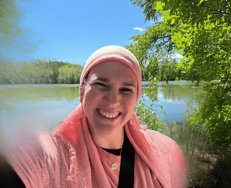 Naomi Korbman smiling wearing a pink headscarf in a selfie by a calm lake with lush green trees.