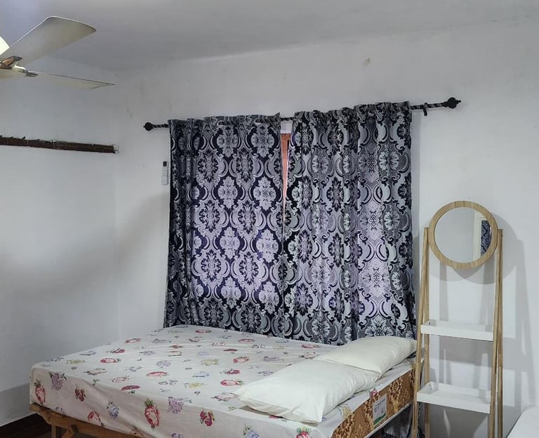 A small bedroom featuring a bed with patterned sheets, blue curtains, a ceiling fan, and a mirror.
