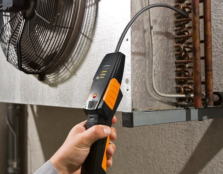 A hand holding a handheld electronic refrigerant leak detector near a freezer unit condenser coil.