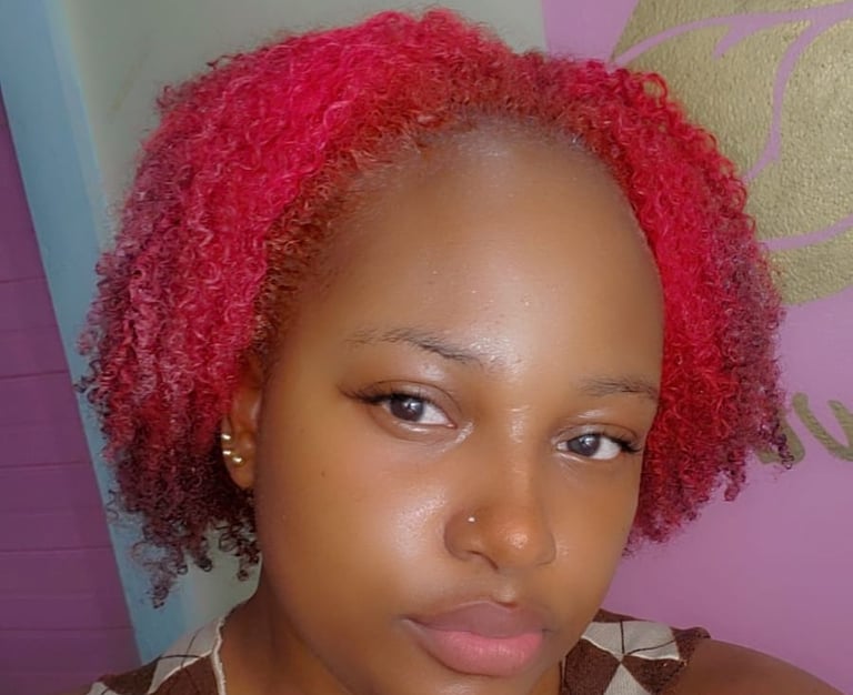 A young woman with rosy pink natural curls, styled in a short, voluminous shape. Her glowing skin, subtle makeup, and nose pi