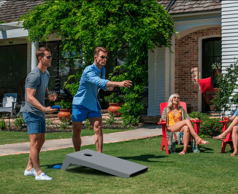 Žmonės žaidžia cornhole žaidimą kieme. Meta žaidimo maišelį į lentą