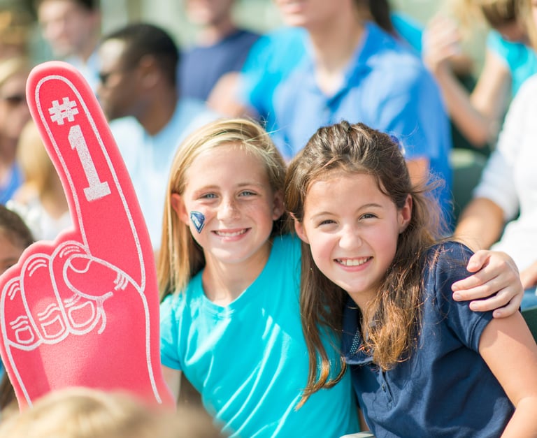 young girls in the stands of athletic event cheering on their team with a foam finger and big smiles