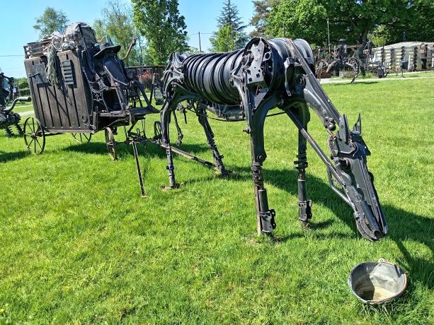 A life-sized steampunk horse sculpture made of scrap metal pulling a rustic carriage on green grass.