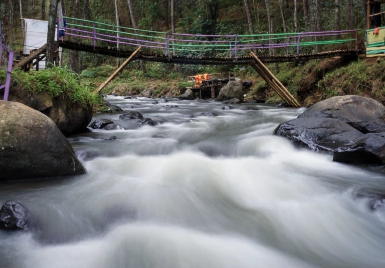 Sebuah jembatan bambu dengan pagar ungu dan hijau membentang di atas aliran sungai deras yang dikelilingi batu besar dan pepo