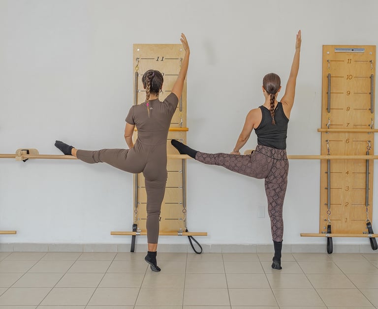 Two women performing a side leg raise stretch against the barre in a Pilates class.