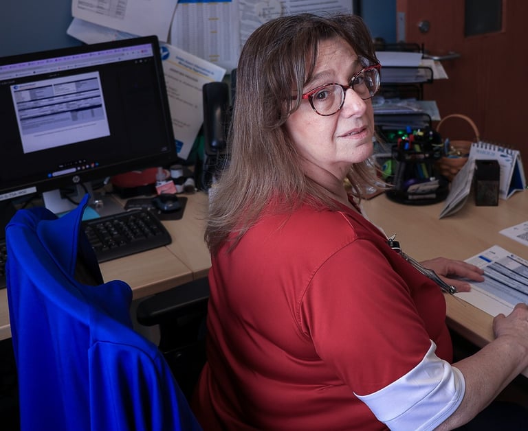 A female office professional wearing glasses and a red shirt reviews documents at her computer desk.