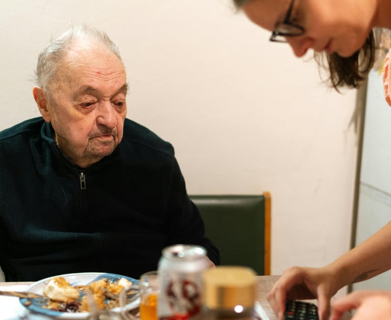 une personne âgée assise autour d'une table regardant son aidante manipuler son portable.