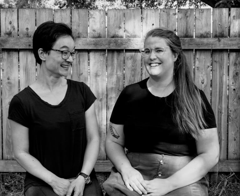 Andy and Jen laughing together while seated outdoors in dappled sunlight in front of a fence.