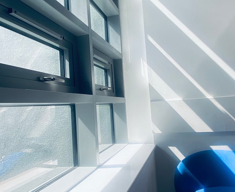Modern office window with natural sunlight shadows casting over a blue chair and white wall.