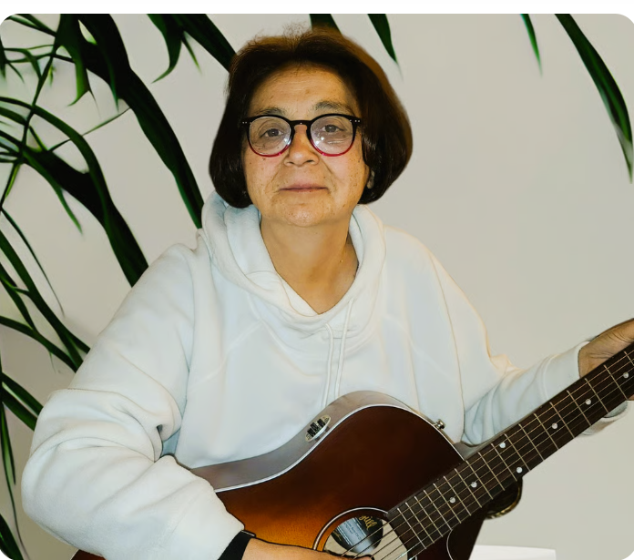 Diane Fischer playing an acoustic guitar indoors next to a green plant.