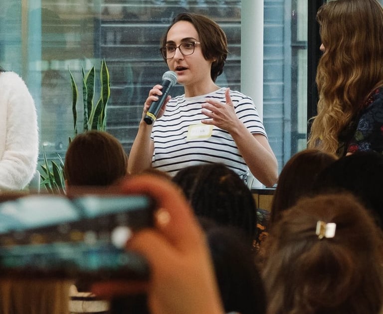 Photo of Micha in a stripy t-shirt holding a microphone and speaking to an audience