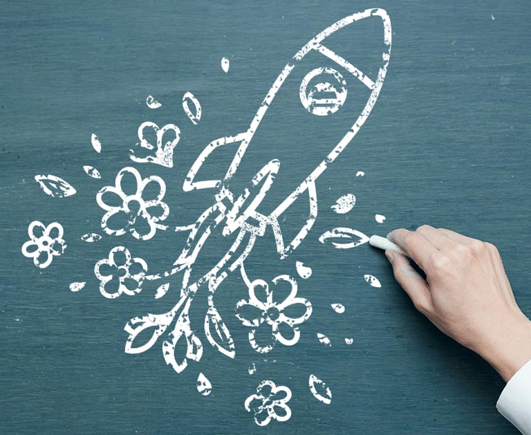 A hand holding white chalk draws a rocket propelled by flowers on a chalkboard.