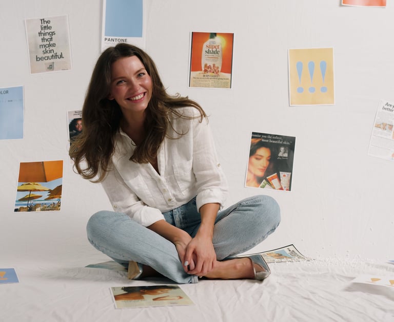 Smiling woman in a white linen shirt and jeans sitting in front of a wall with skincare mood board inspiration.