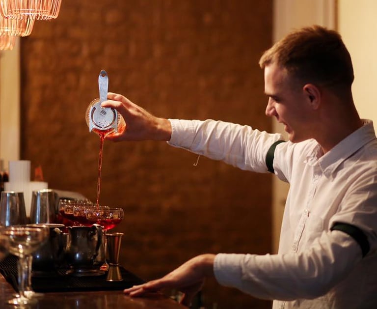 Professional bartender straining a red craft cocktail into glasses at a dimly lit upscale bar.