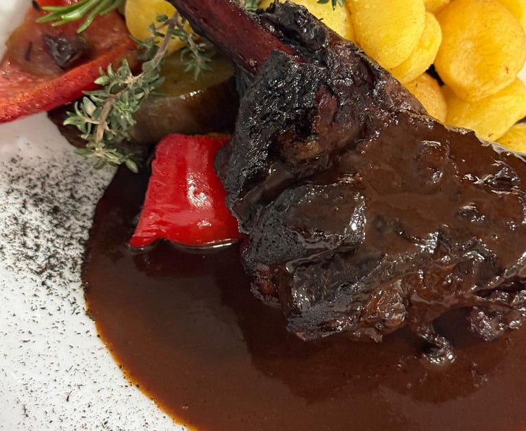 Braised lamb shank with rich red wine sauce, roasted potatoes, and fresh rosemary garnish.