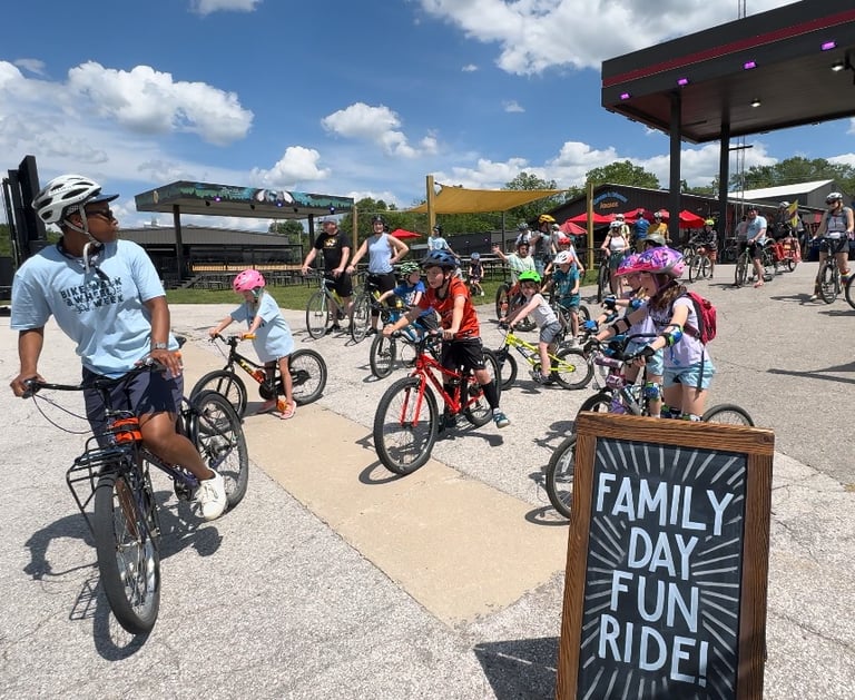 A group of children on bikes, led by an adult, right behind a sign reading "Family Day Fun Ride"