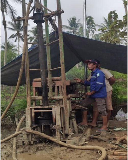 A worker operates a portable water well drilling rig at an outdoor construction site.