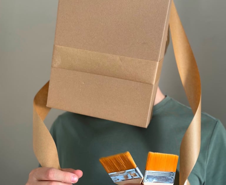 Photo shows a man with cardboard boas on his head covering his face. Man holds paint brushes.