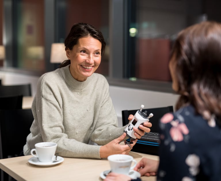 Woman in cafe, holding an iCare HOME2 and discussing with her friend, about measuring her own eye pressure IOP at home.