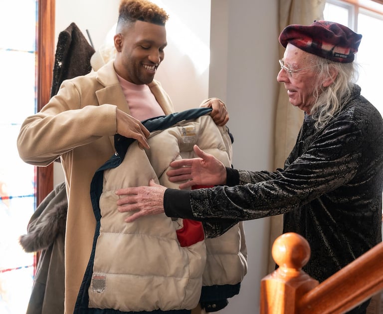 A young man in a tan overcoat helps a senior man put on a winter puffer jacket at home.