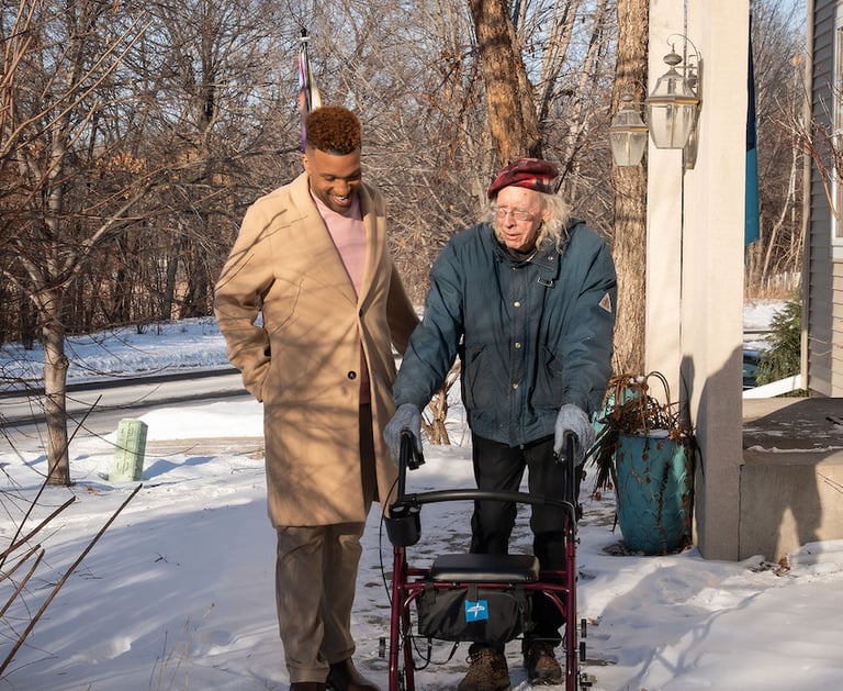 A young caregiver assists a senior man using a walker on a snowy sidewalk outside a home.