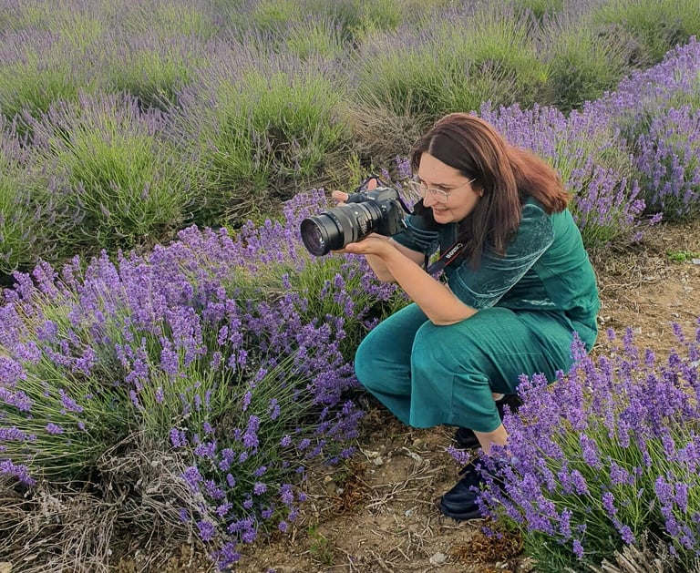 A female photographer holding a camera crouches in a blooming purple lavender field.