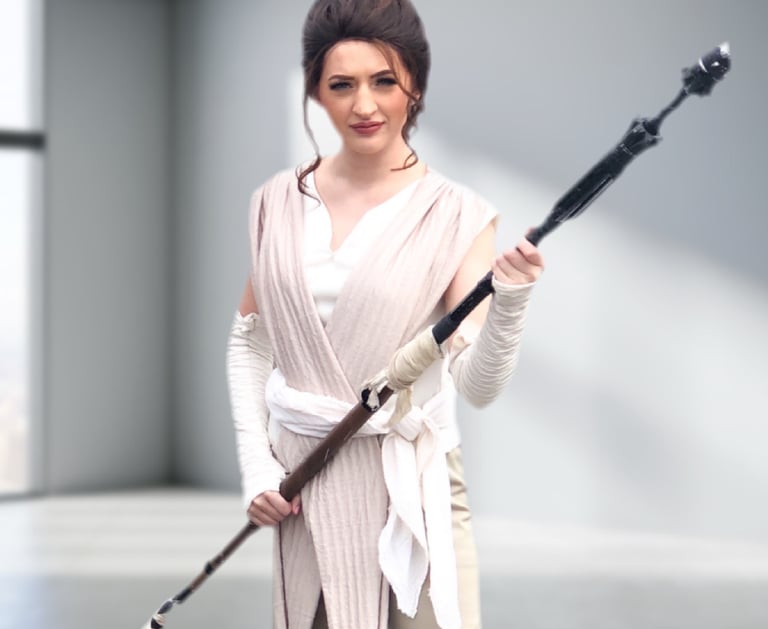 A woman in a realistic Rey Skywalker cosplay holding a staff in a minimalist studio.