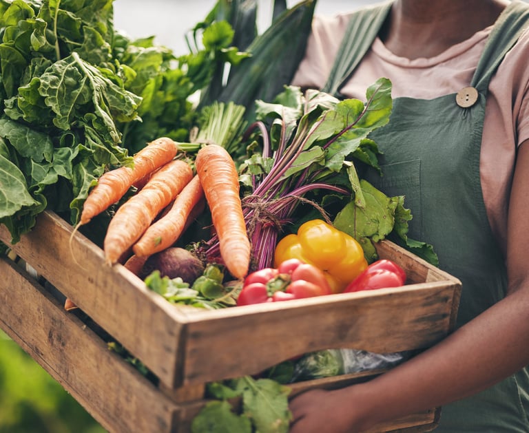 A farmer holding a wooden crate filled with fresh organic vegetables like carrots, peppers, and leafy greens.