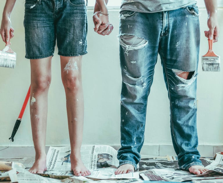 A couple in paint-splattered jeans holding hands and brushes during a DIY home renovation.