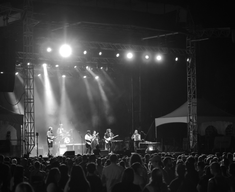 A live band performs on a brightly lit outdoor stage for a large crowd at a night concert festival.